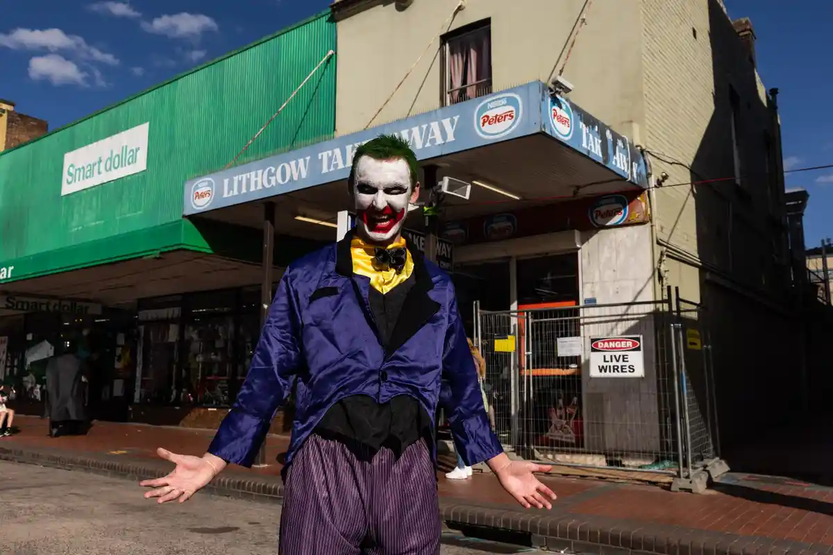 A person dressed as the Joker from Batman, with white face paint, red lips, green hair, and a purple suit, stands on a…