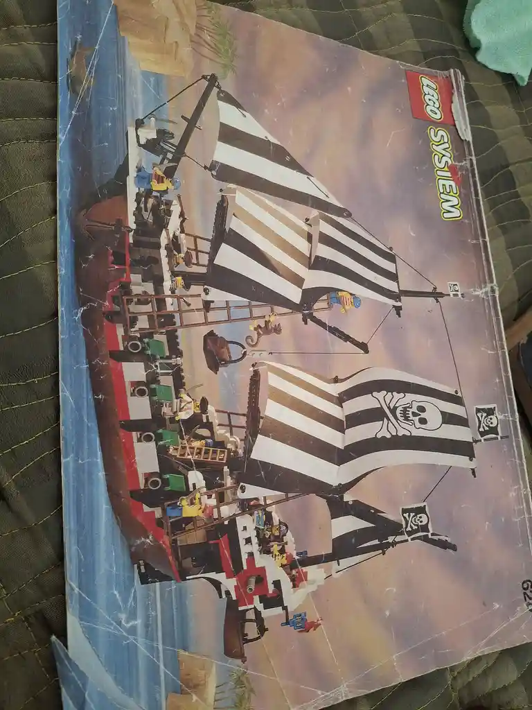 A weathered, scratched LEGO System box depicts a large pirate ship made of LEGO bricks with black-and-white striped sails…