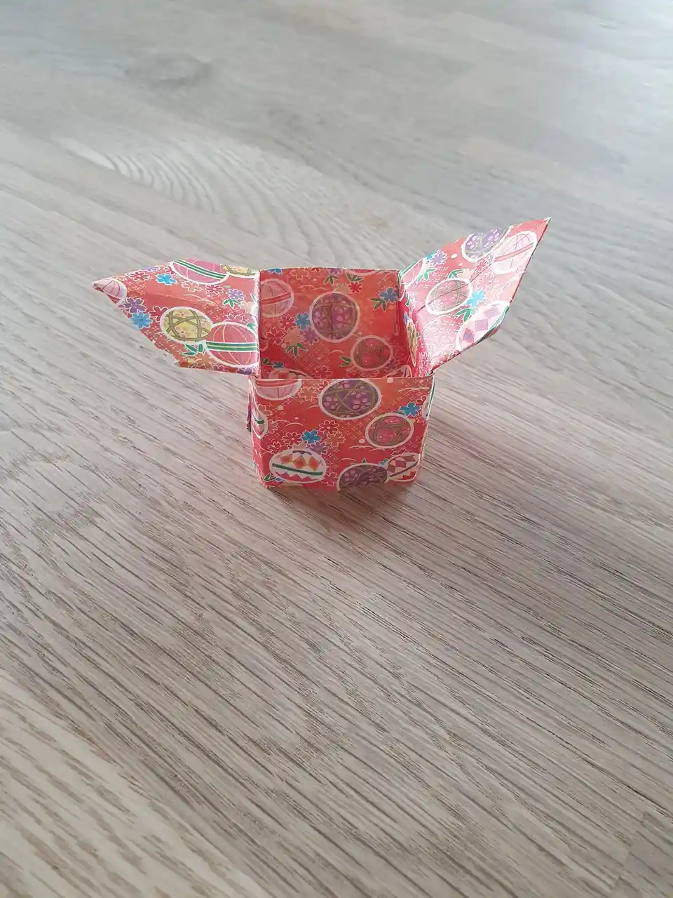 A small, colorful origami box with a red-orange pattern of flowers and circular designs sits open on a light wood-grain…