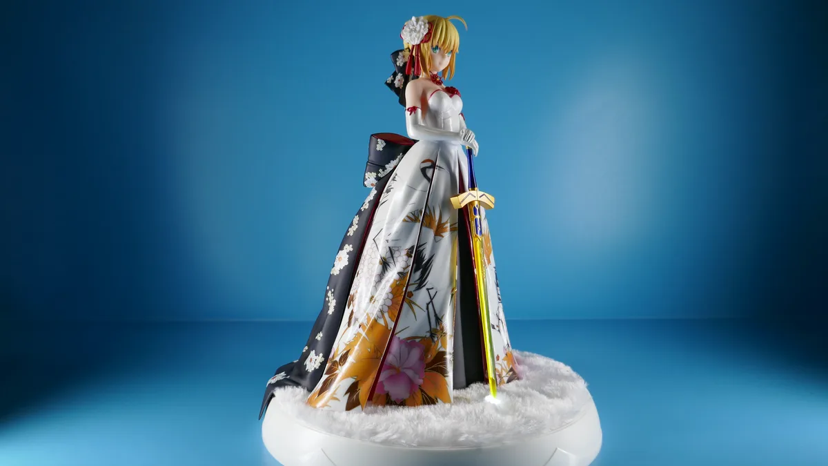 A figurine of a blonde anime-style woman in a white wedding dress with floral patterns and a black obi, holding a sword…