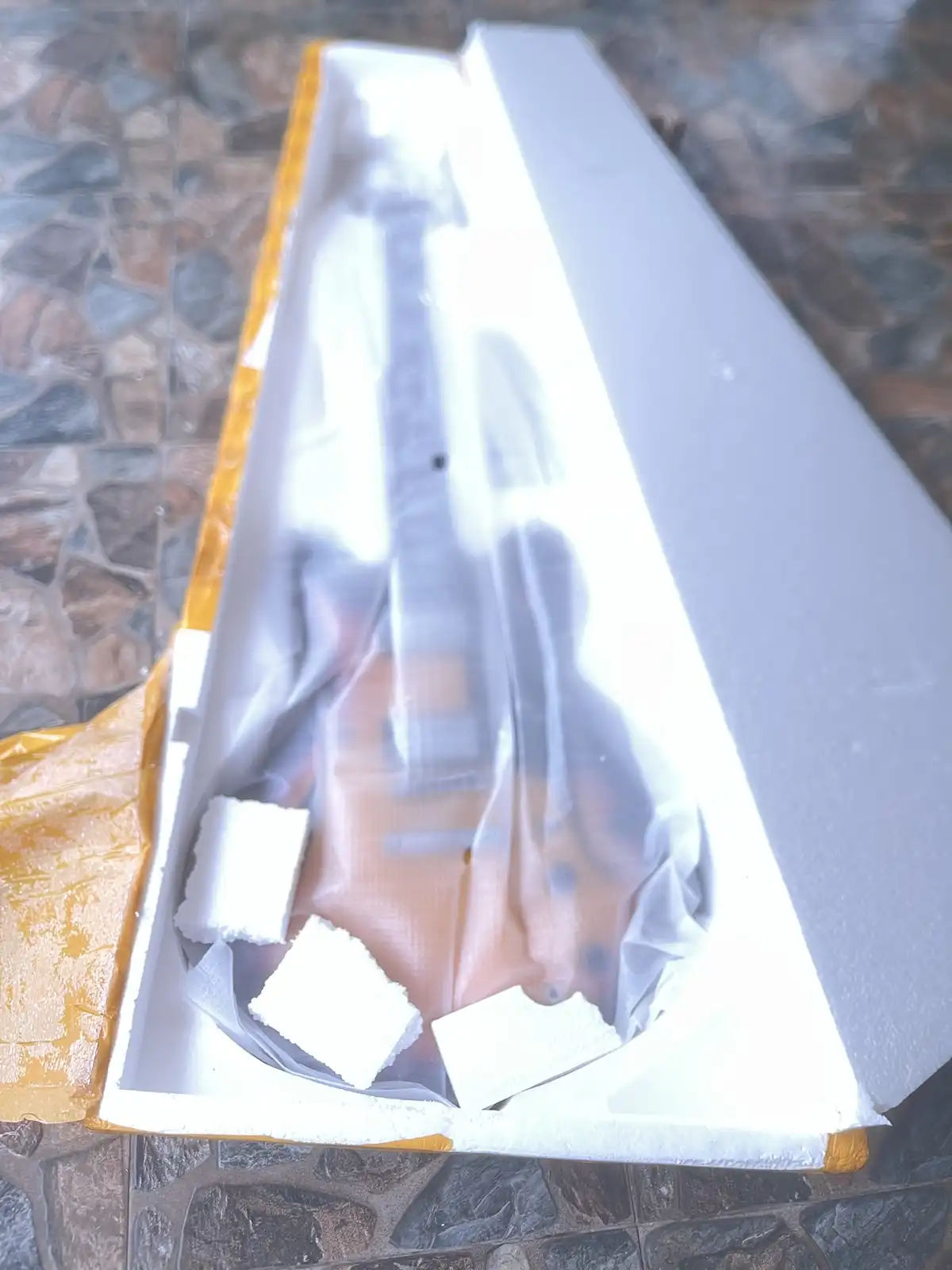 A long, flat cardboard box with yellow tape, opened to reveal a guitar wrapped in clear plastic inside, resting on white…