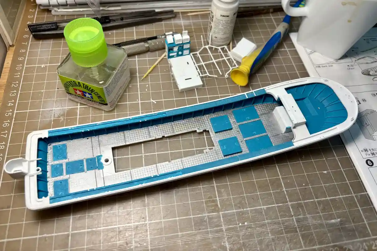 A partially assembled model ship hull lies on a gridded work mat, its interior painted blue while the exterior remains…