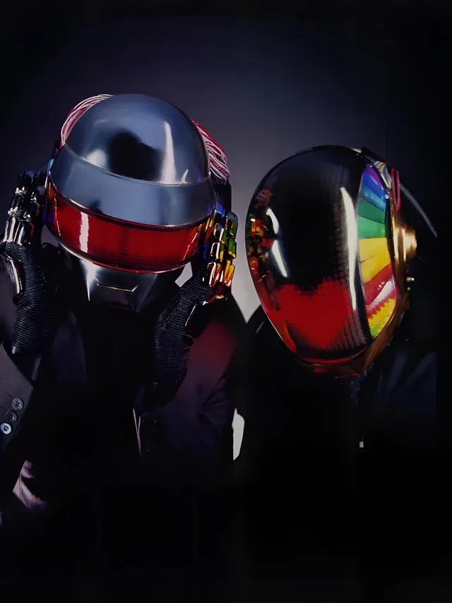 Two figures in iconic Daft Punk helmets, one with a red visor and the other with a rainbow-colored visor, stand side by side…