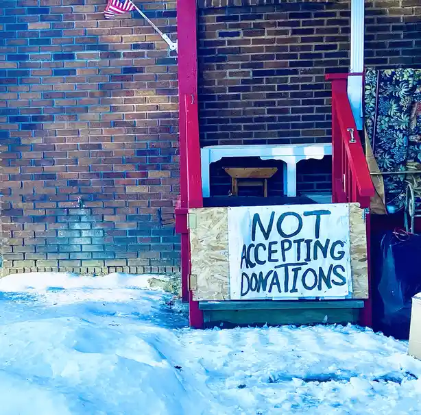 A snowy outdoor scene shows a makeshift donation box constructed from plywood and a white table, placed at the base of…