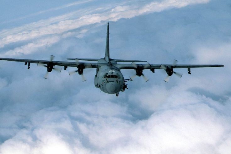 A military transport aircraft, likely a C-130 Hercules, is shown flying directly toward the viewer above a layer of clouds…
