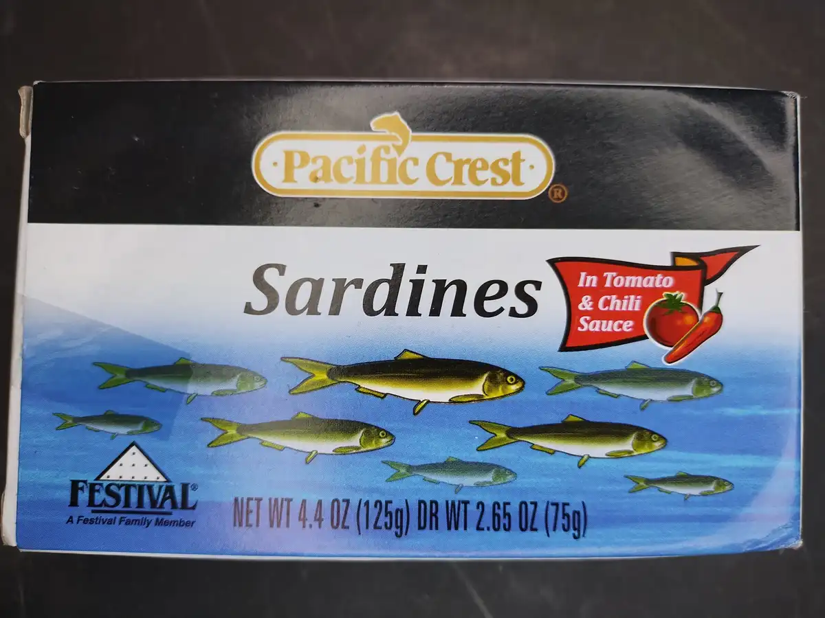 A close-up of a Pacific Crest sardines box, showing illustrated fish swimming in blue water, with text indicating 'In Tomato…