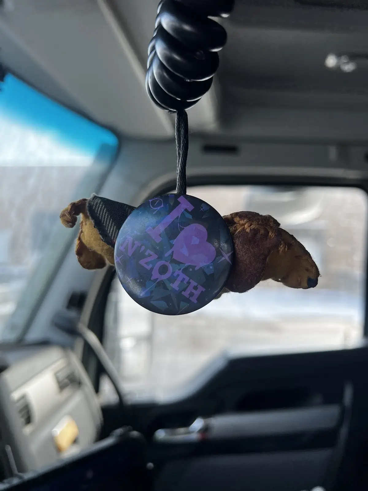 A close-up shot from inside a car shows a small, brown plush dog hanging from a black coiled cord attached to the rearview…