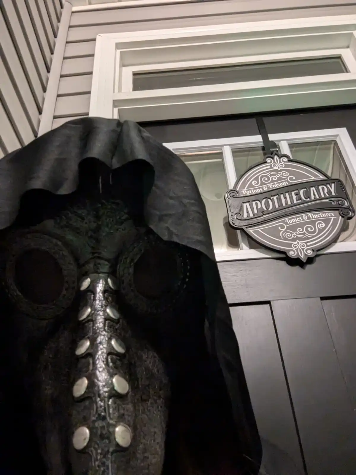 A close-up photo shows a large, ornate plague doctor mask with a black hood and silver buttons, positioned in front of a…
