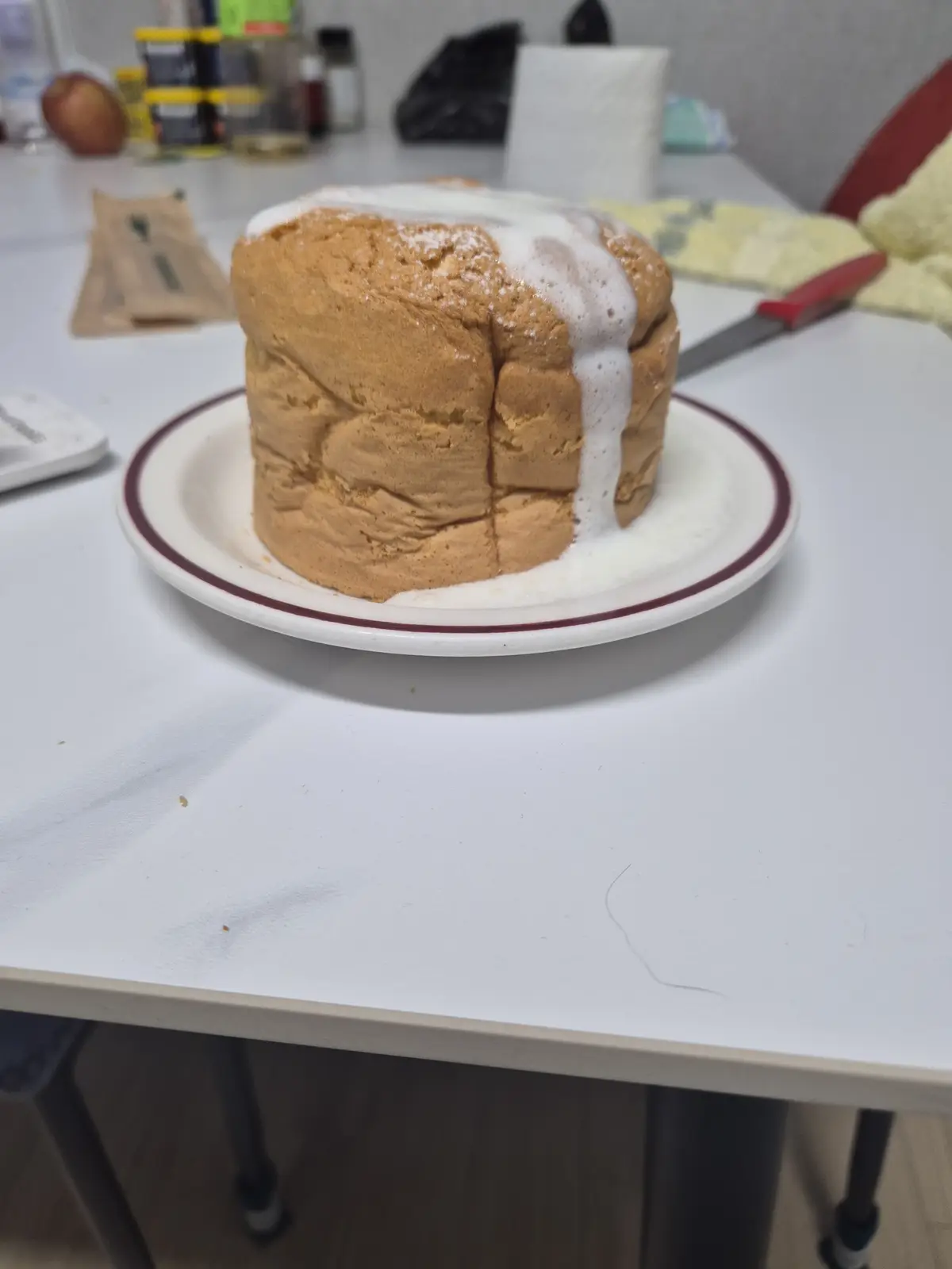 A tall, golden-brown cake sits on a white plate with a red rim, covered in a thick, white, foamy substance that drips down…