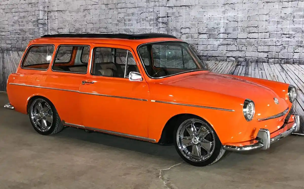 A bright orange vintage station wagon with chrome wheels and trim is parked indoors against a gray brick wall. The car has a…