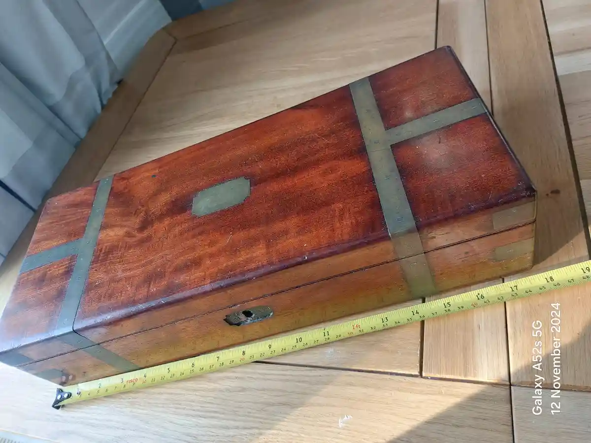 A long, rectangular wooden box with brass straps and a metal latch sits on a light wood table, with a yellow measuring tape…