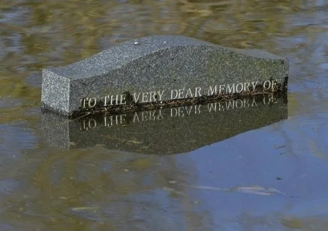A granite gravestone partially submerged in murky water, with its engraved text 'TO THE VERY DEAR MEMORY OF' visible above…