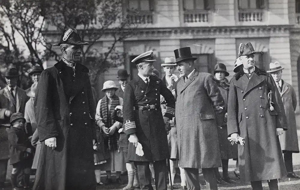 A black-and-white historical photo shows four men in formal early 20th-century attire standing outdoors in front of a…