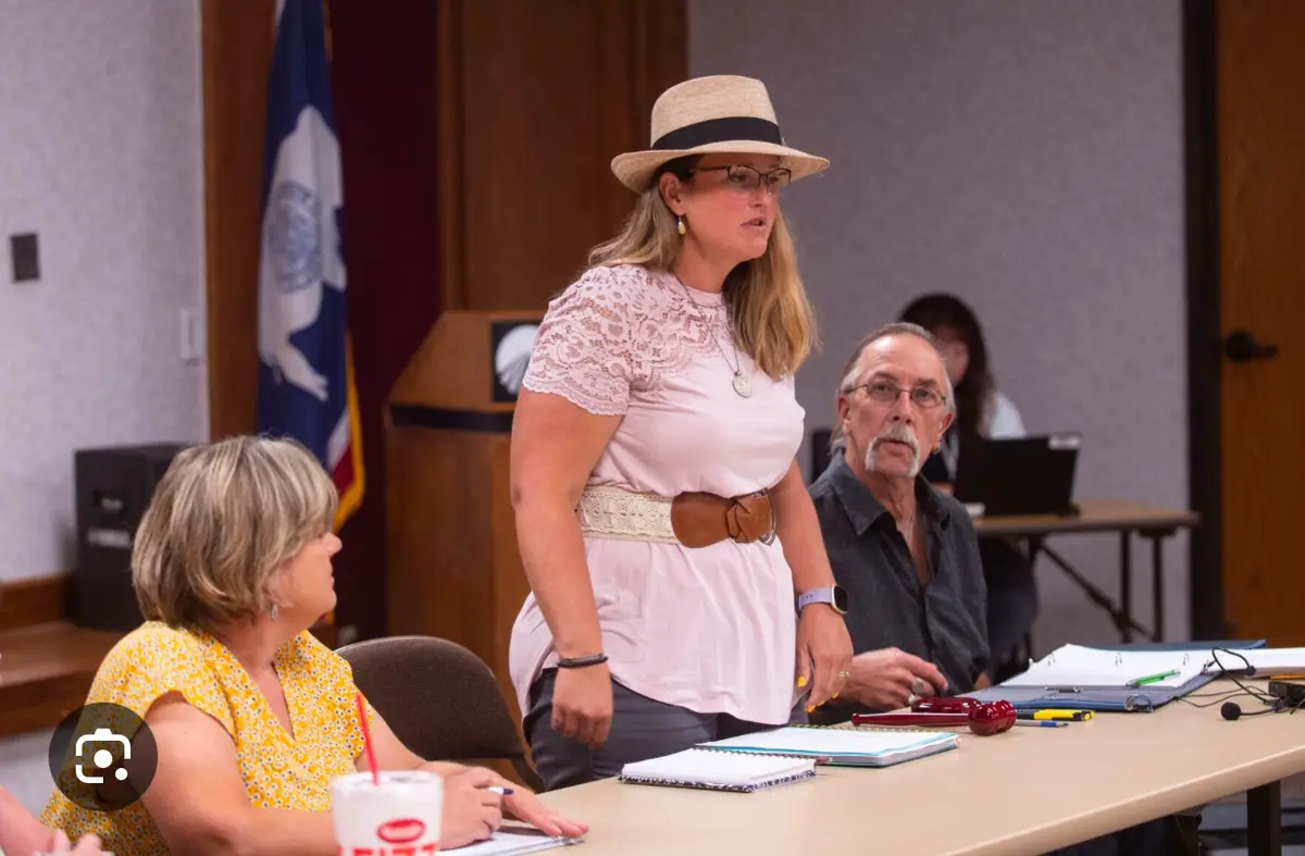 A woman in a straw hat and glasses stands at a conference table in a formal meeting room, speaking while others sit around…