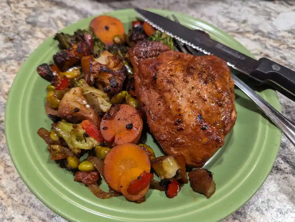 A close-up photo of a green plate holding a grilled chicken breast and a colorful mix of roasted vegetables including…