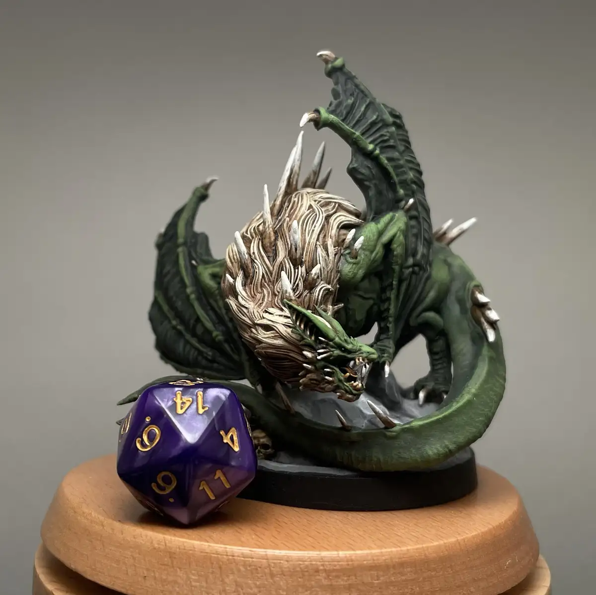 A green dragon miniature with spiked wings and a detailed, textured head is posed on a wooden display stand. Beside it rests…