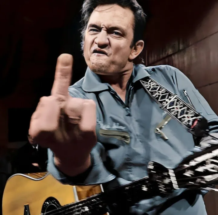 a man with dark hair and a light blue shirt is holding an acoustic guitar with a patterned strap, pointing his right index…