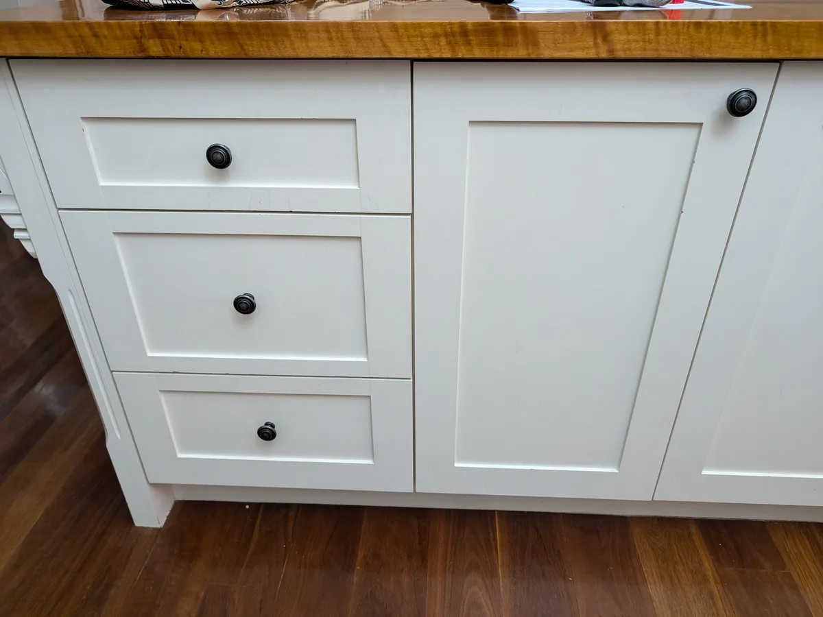 A close-up photo of white kitchen cabinets with a wooden countertop and dark wood flooring. The left side has three stacked…