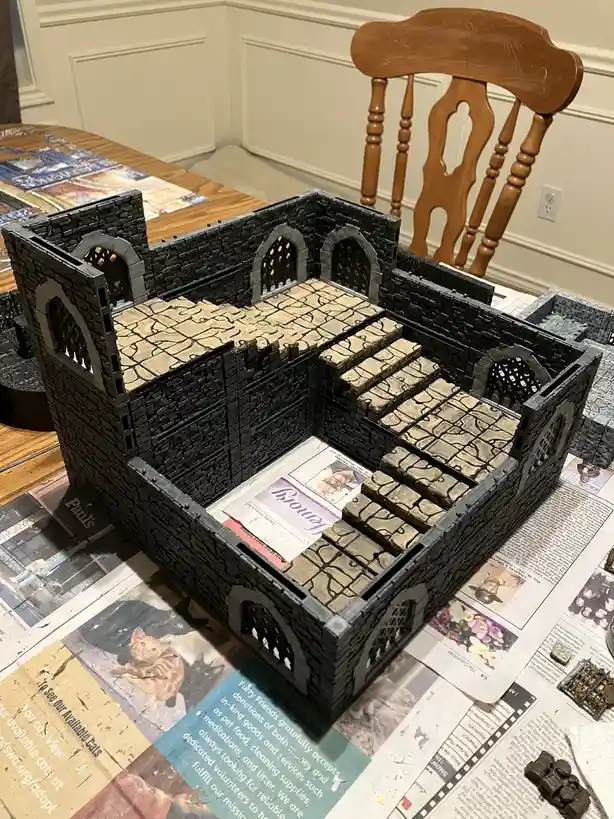 A 3D-printed, castle-like structure made of black and beige plastic pieces sits on a table covered with newspaper, with a…