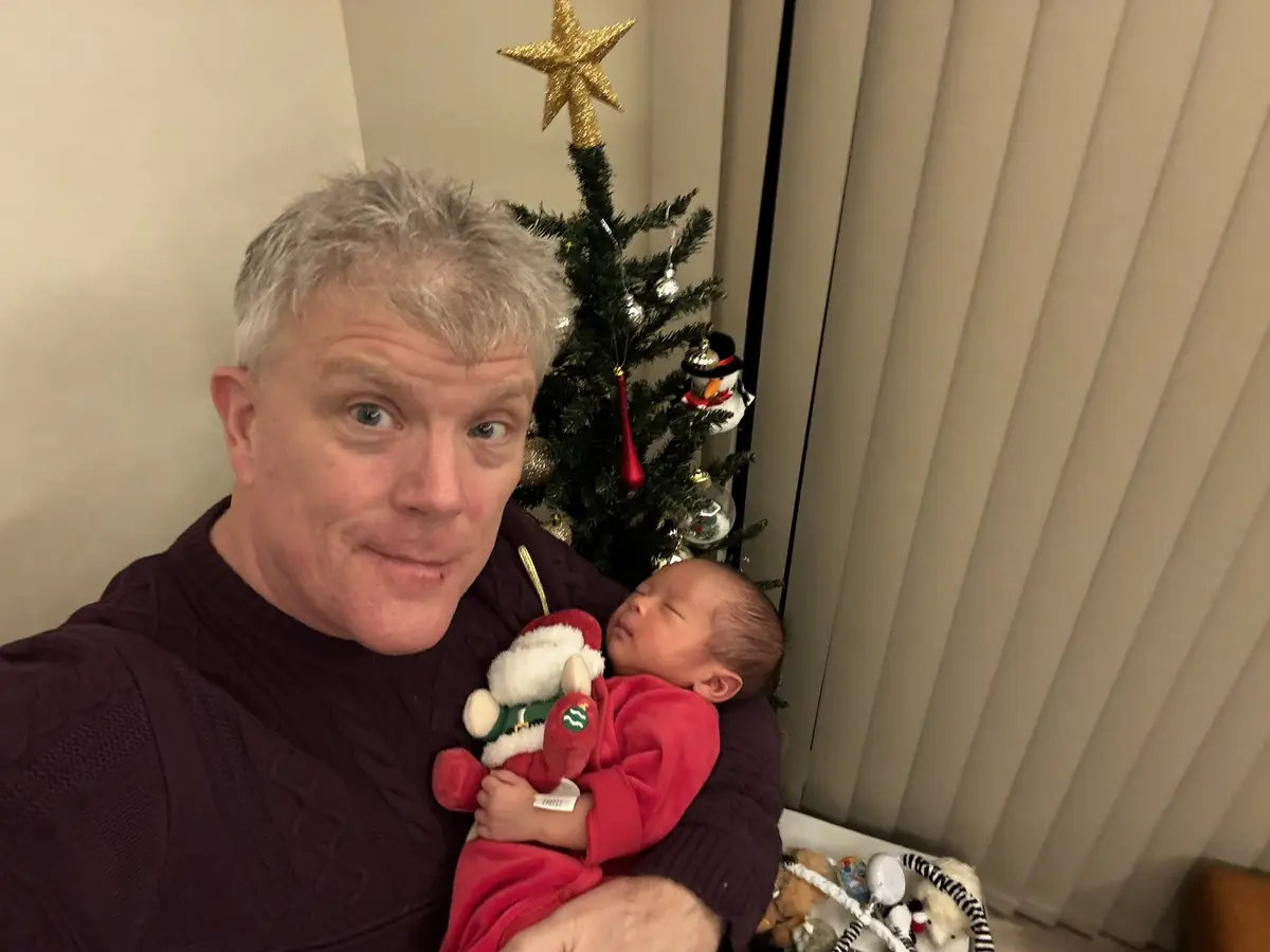 A man with gray hair wearing a dark sweater holds a sleeping newborn baby dressed in red, who clutches a small Santa plush…