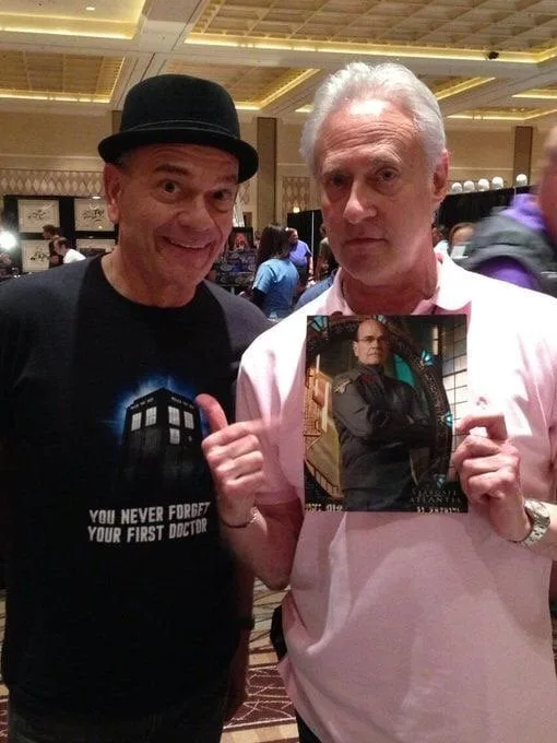 Two men pose for a photo at a convention. The man on the left, wearing a black t-shirt with a TARDIS graphic and the text…