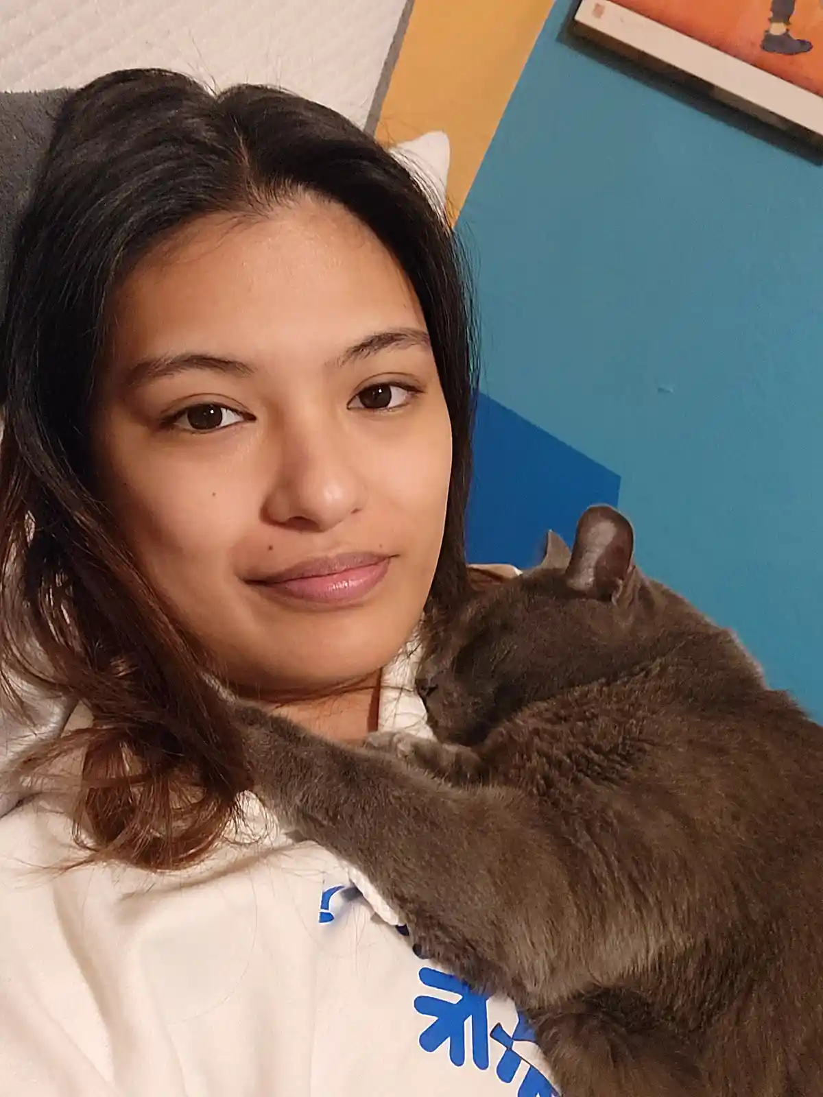 A young woman with dark hair and a slight smile looks at the camera while a gray cat sleeps curled up on her shoulder. She…