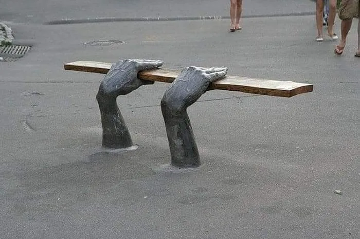 A public bench with a wooden seat is supported by two large, gray, sculpted hands emerging from the pavement, palms up…