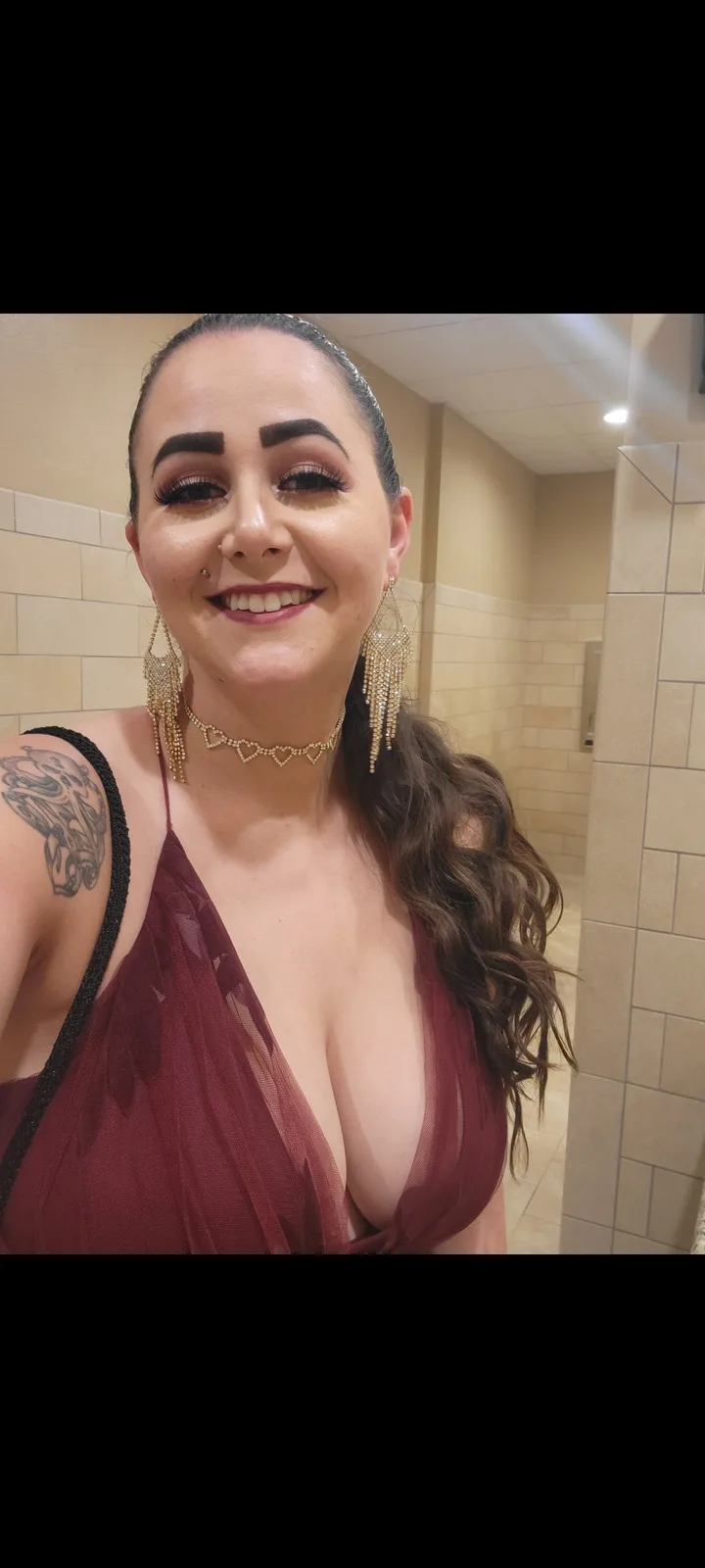 A woman in a deep red dress with heavy makeup, including thick, perfectly arched eyebrows, smiles at the camera in a tiled…