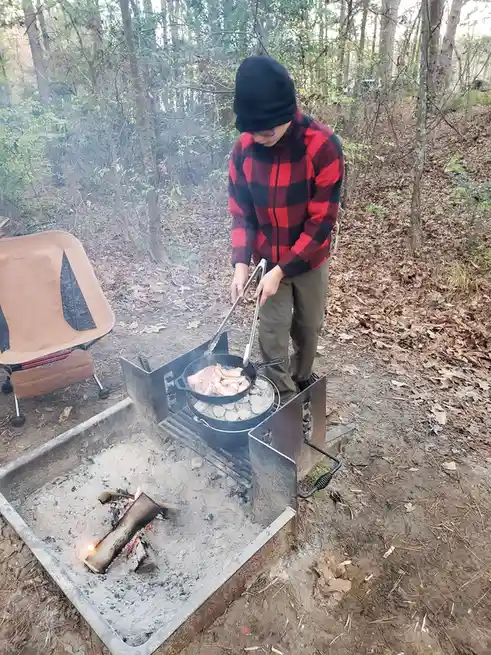 A person in a red and black plaid jacket and black beanie is cooking bacon in a cast iron skillet over a campfire in a…
