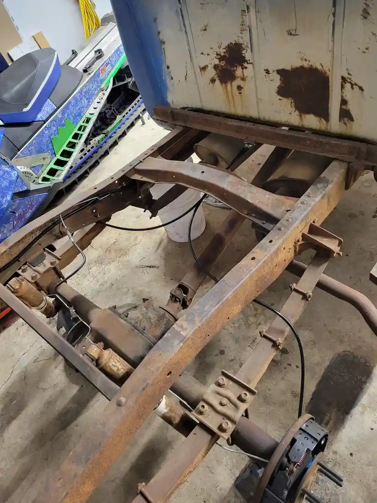A rusty, stripped-down vehicle chassis sits in a garage, showing exposed metal frames, bolts, and wires. In the background…