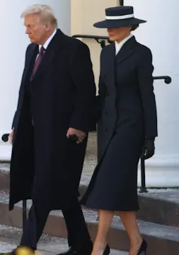 A man and a woman in formal dark attire walk side by side outdoors. The woman wears a black hat with a white band and a…