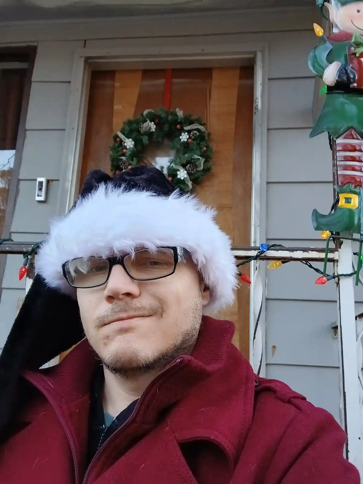 A man wearing a black and white furry Santa hat and glasses stands in front of a house decorated for Christmas with a wreath…