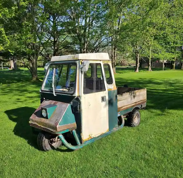 a weathered, rusty, three-wheeled utility vehicle with a white and teal paint job sits parked on a lush green lawn under…