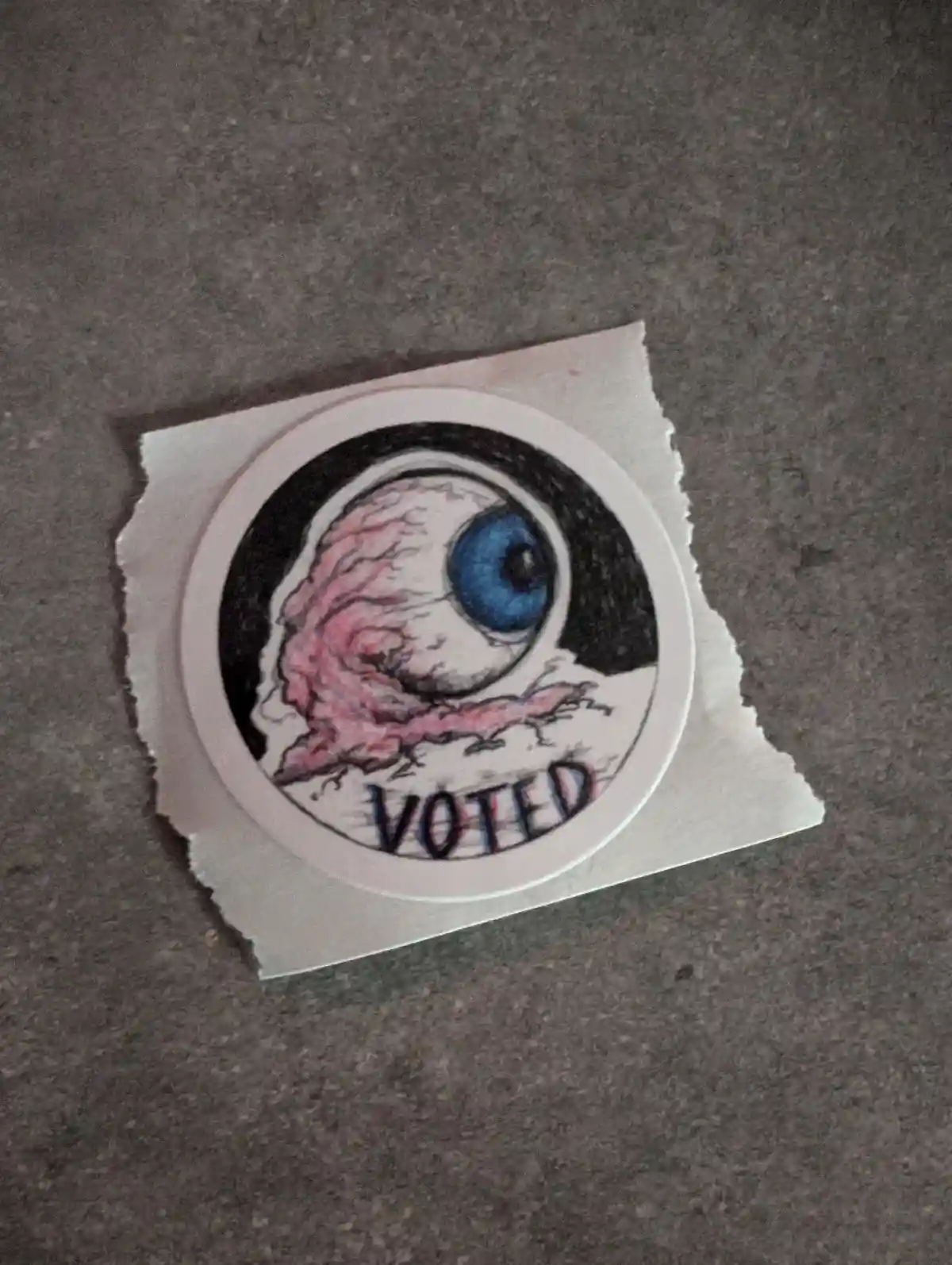 A hand-drawn sticker depicts a grotesque, partially detached eyeball with a blue iris and pinkish, decaying flesh, set…