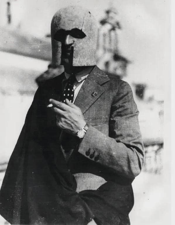 A black-and-white historical photograph shows a man in a suit and tie wearing a large, ancient-style helmet with eye slits…