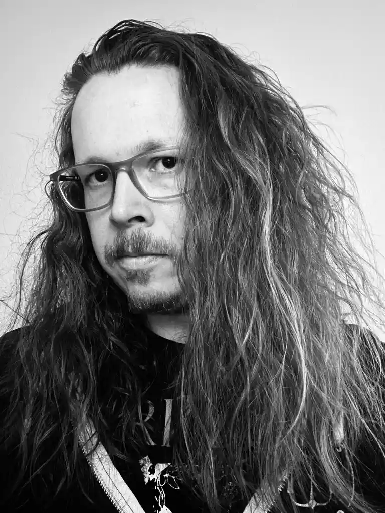 A black-and-white portrait of a man with long, wavy hair, glasses, and a goatee, looking directly at the camera with a…