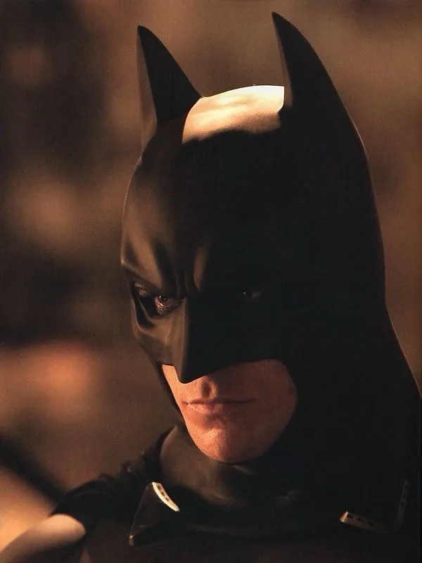 a close-up of the character Batman from the 1997 film, wearing his iconic black cowl with pointed ears and a white bat…