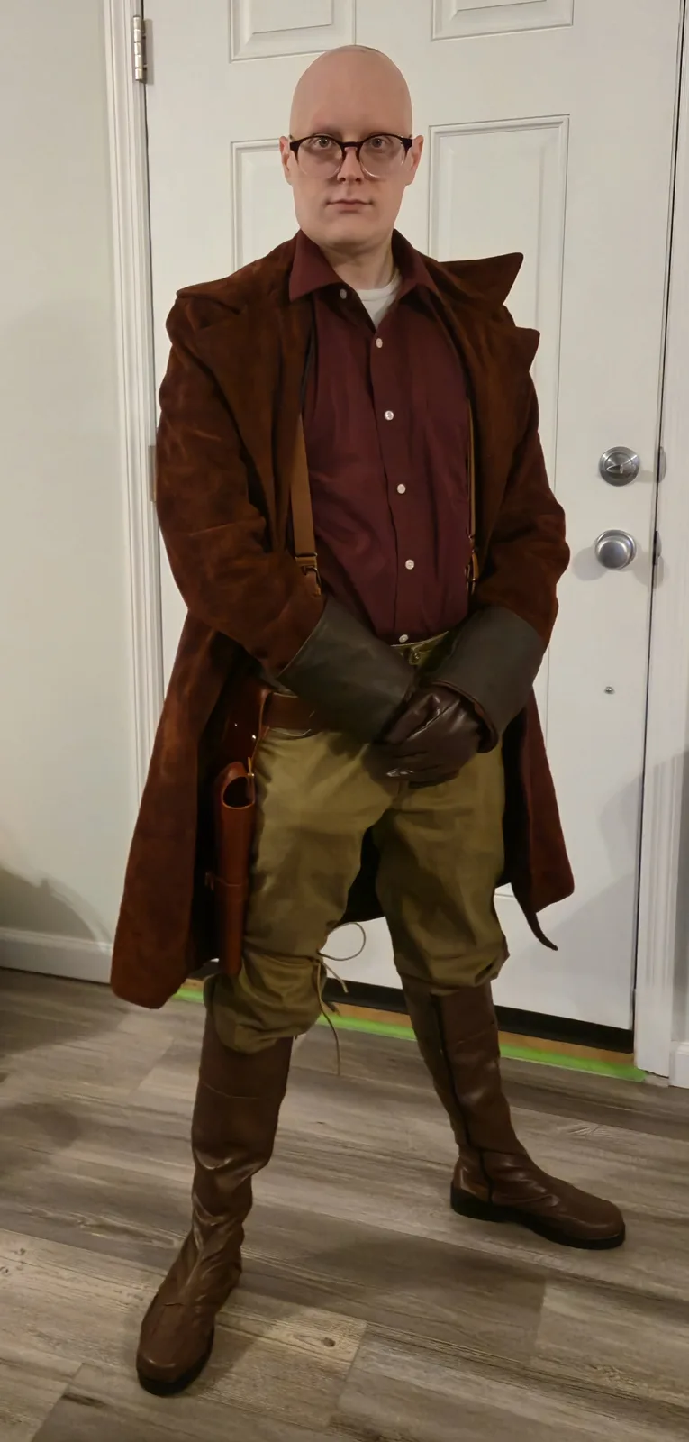 A bald man wearing glasses stands indoors in a full fantasy costume featuring a brown suede coat, maroon shirt, olive pants…