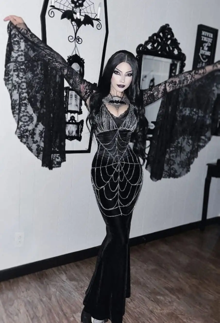 A woman in a black, spiderweb-patterned velvet dress with lace sleeves poses with arms outstretched, mimicking bat wings…