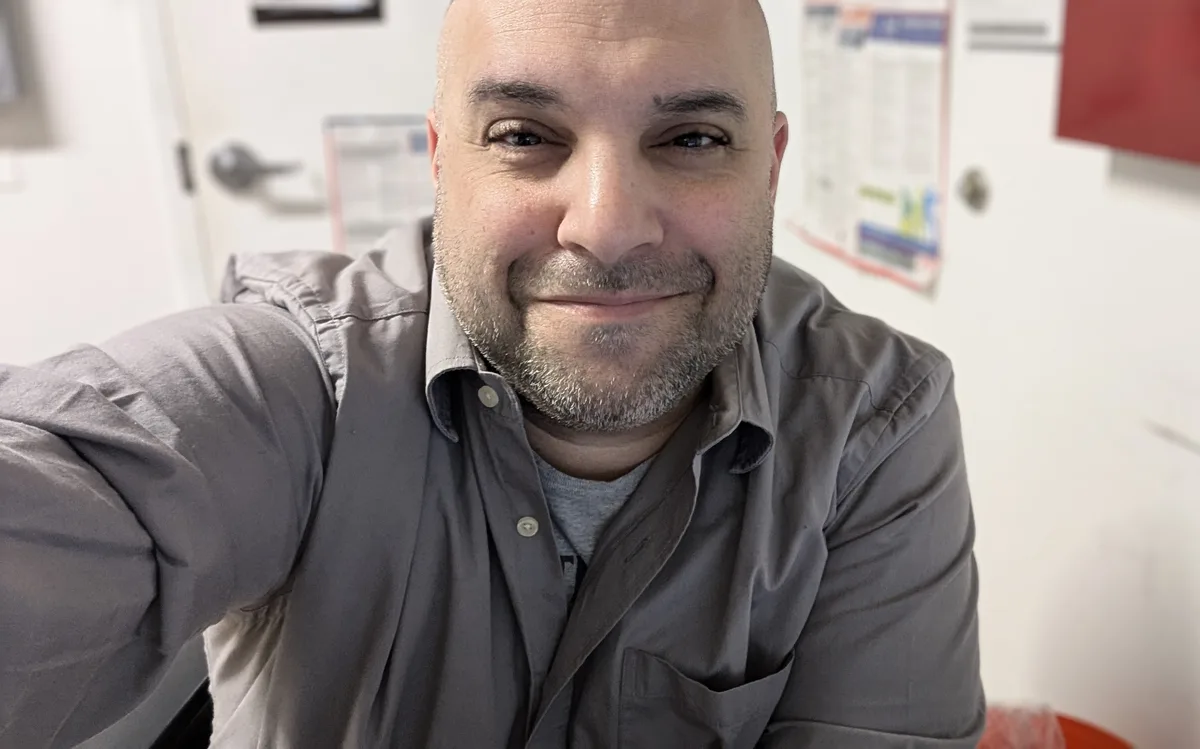 A bald man with a gray beard and mustache, wearing a gray button-up shirt over a gray t-shirt, takes a selfie in what…