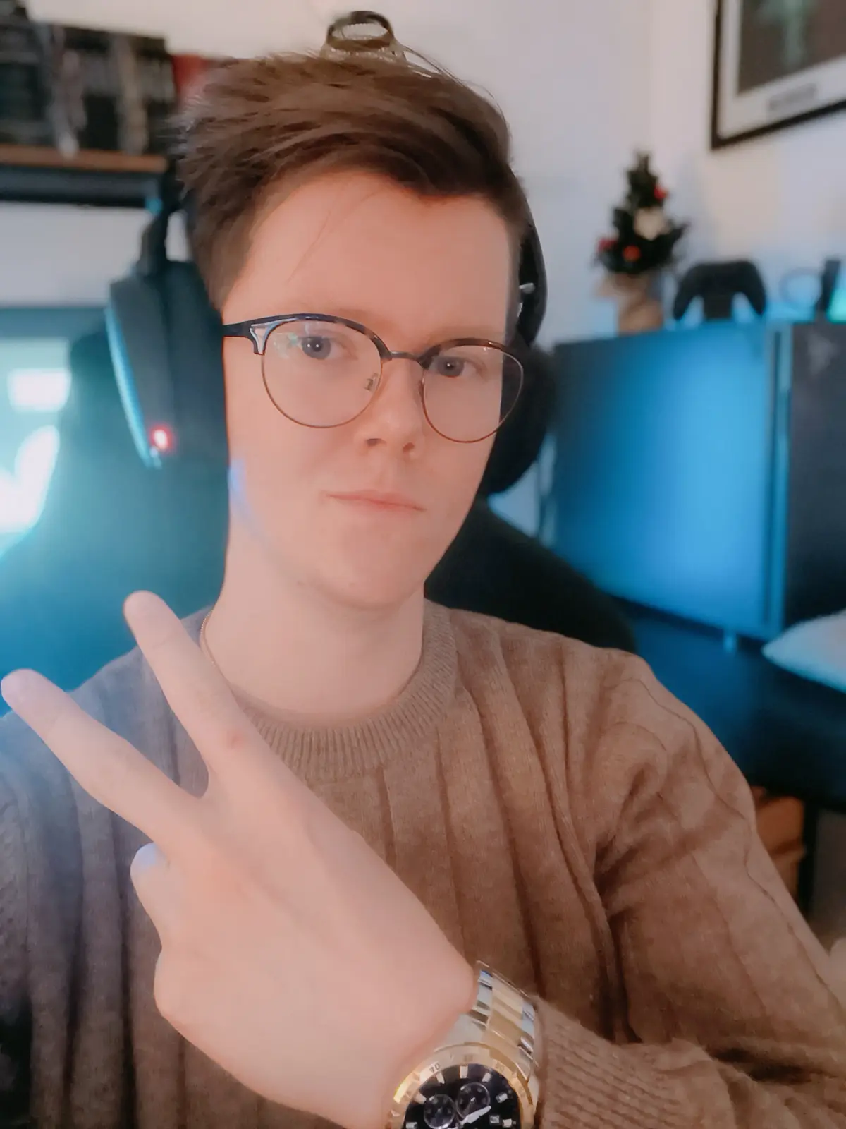 A young man with glasses and headphones makes a peace sign with his hand while wearing a gold watch, looking directly at the…