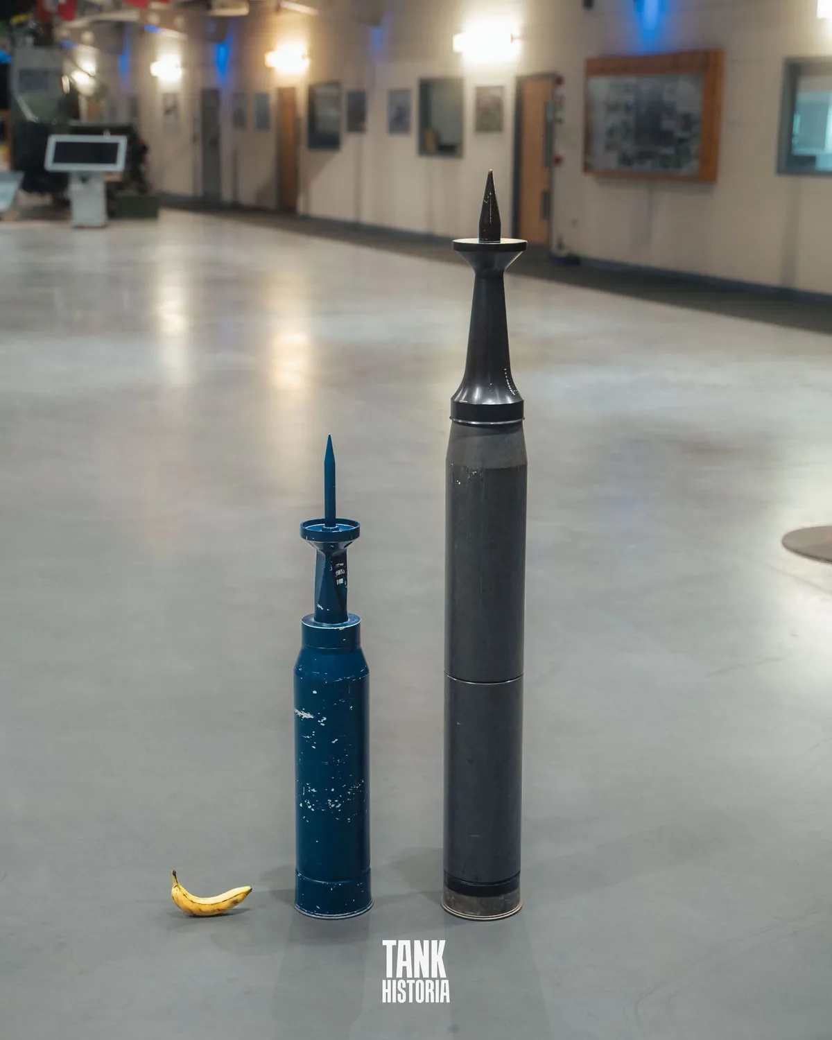 Two large, pointed tank shells stand upright on a polished floor next to a single banana, creating a humorous size…