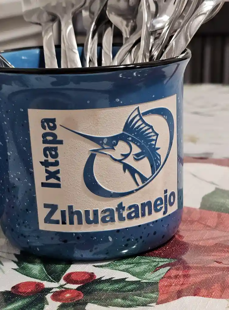 A blue speckled enamelware utensil holder filled with silver forks, featuring a logo of a marlin and the text 'Ixtapa…