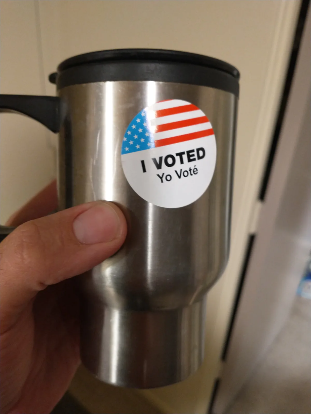 A hand holds a stainless steel travel mug with a black lid and handle. Stuck to the side is a circular sticker featuring the…