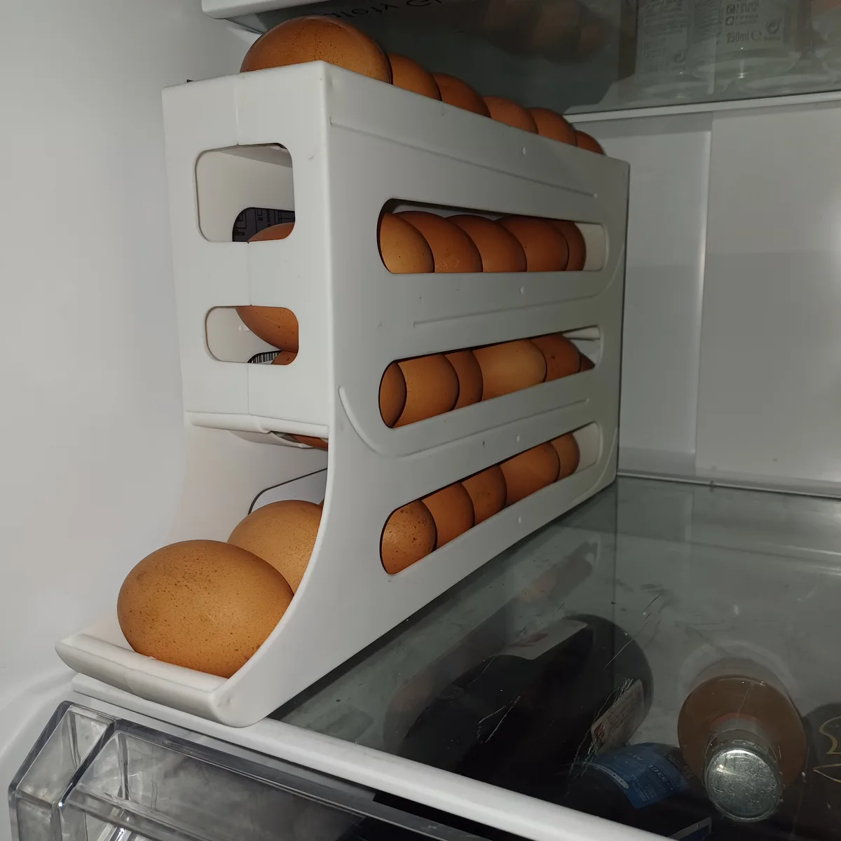 A white plastic egg holder inside a refrigerator is filled with numerous brown eggs, with some spilling over the top and a…