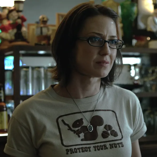 A woman with glasses and short brown hair wears a light-colored t-shirt featuring a graphic of a squirrel holding a nut and…