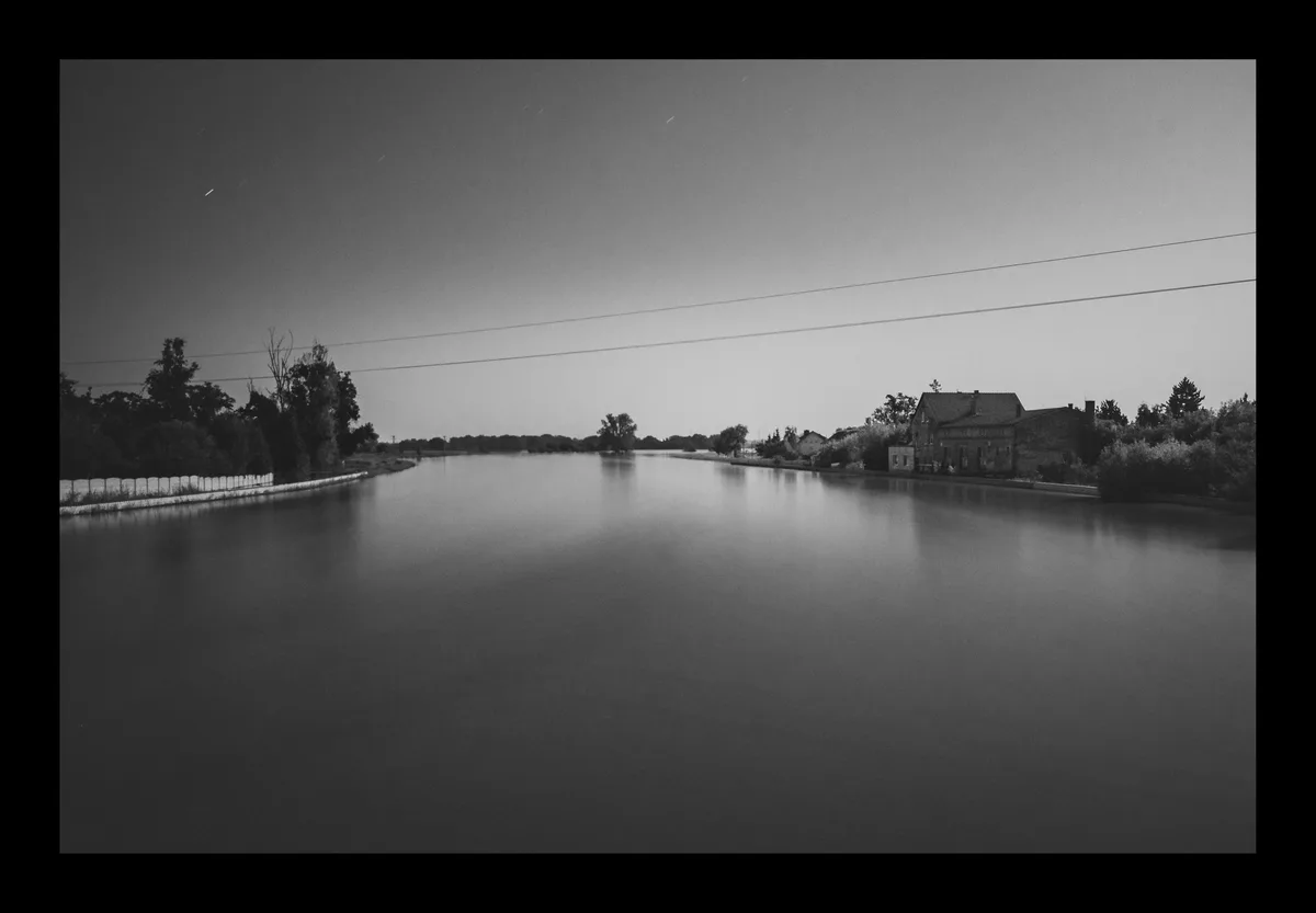 A black-and-white long-exposure photograph of a calm canal at night, showing star trails in the sky, trees, a house on the…