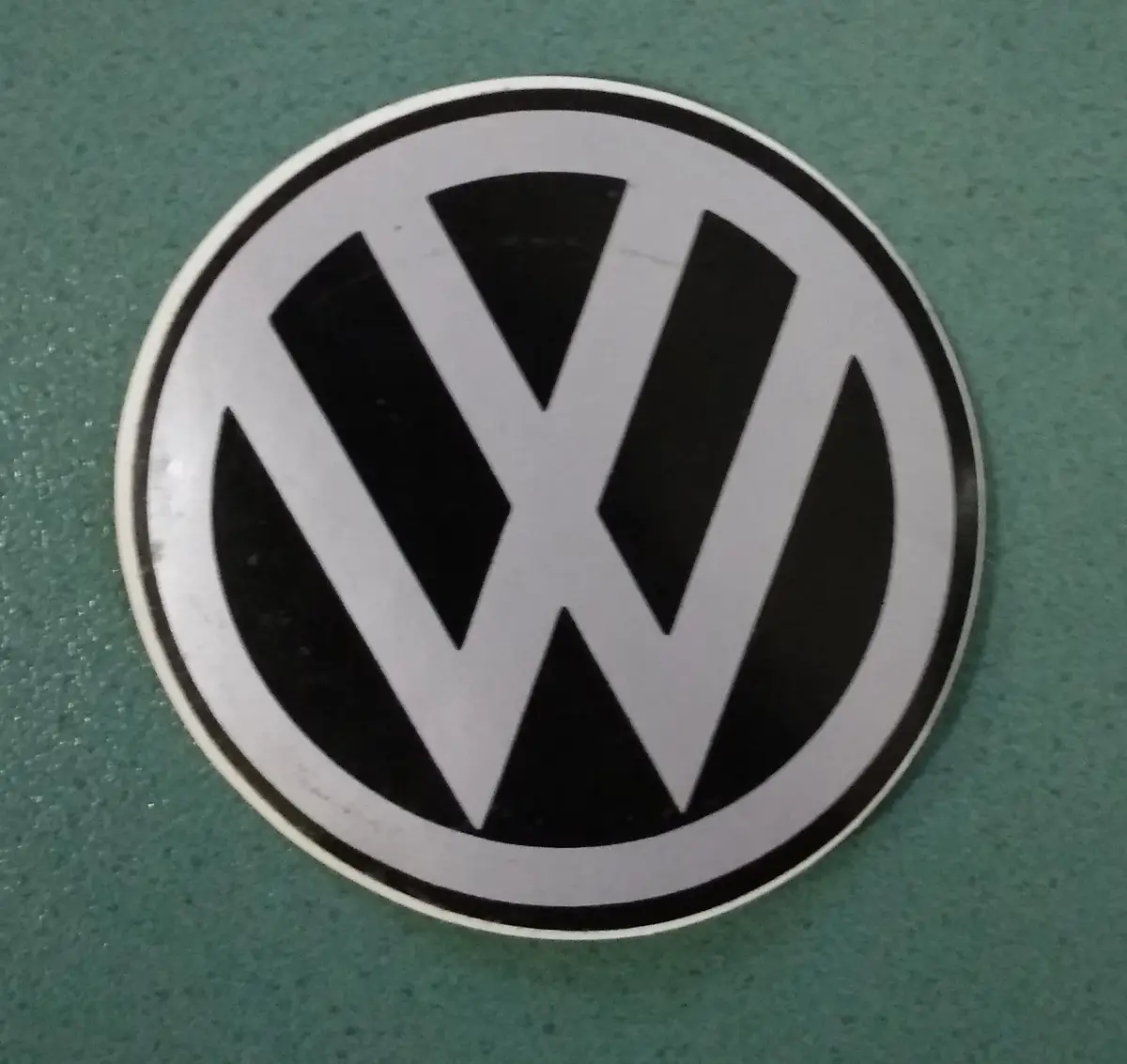 A close-up photo shows a circular Volkswagen logo sticker with a black and white design on a green speckled surface. The…