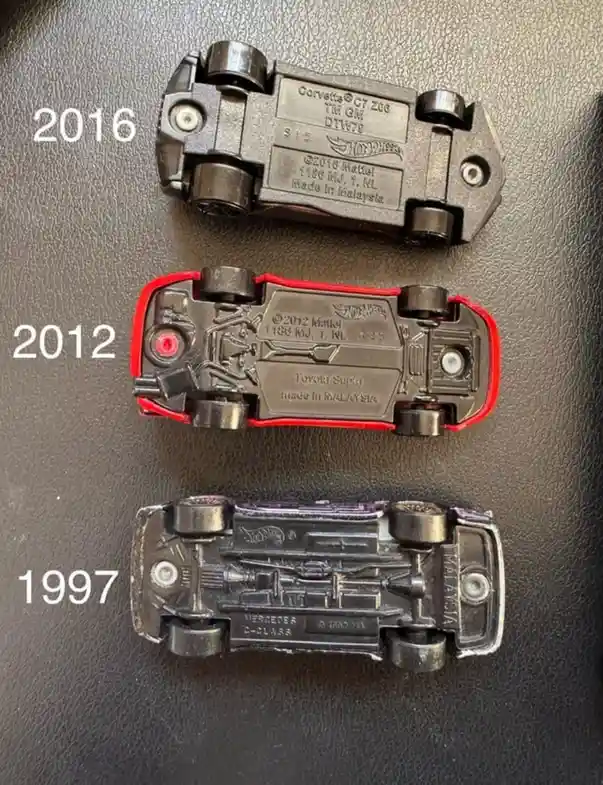 Three Hot Wheels toy cars are shown upside down on a dark textured surface, each labeled with a year: 2016 (Corvette C7…