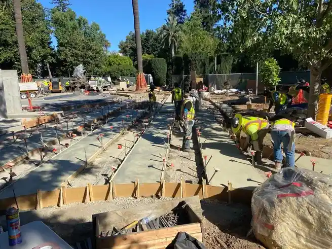 A photo of construction workers laying concrete slabs outdoors under a sunny sky, surrounded by trees and construction…